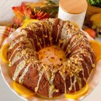 Pumpkin Walnut Bundt Cake on a white cake plate surrounded with orange slices and topped with a delicious orange glaze and chopped walnuts.