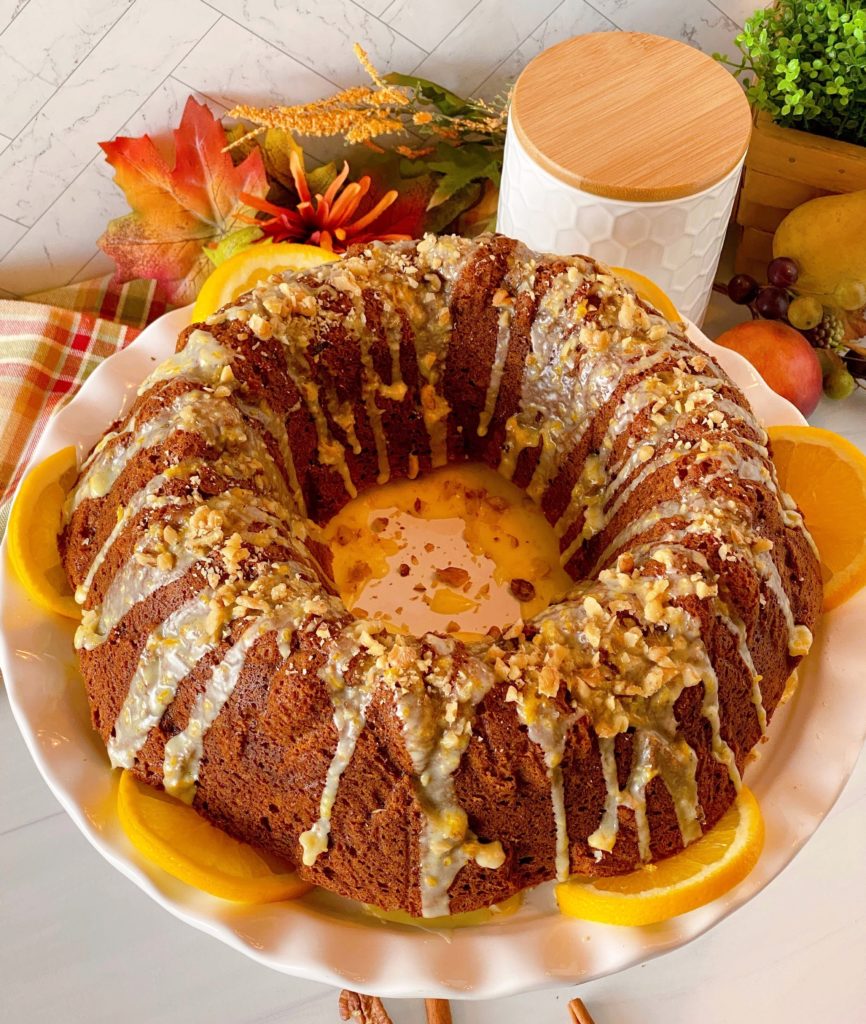 Pumpkin Walnut Bundt Cake on a white cake plate surrounded with orange slices and topped with a delicious orange glaze and chopped walnuts.