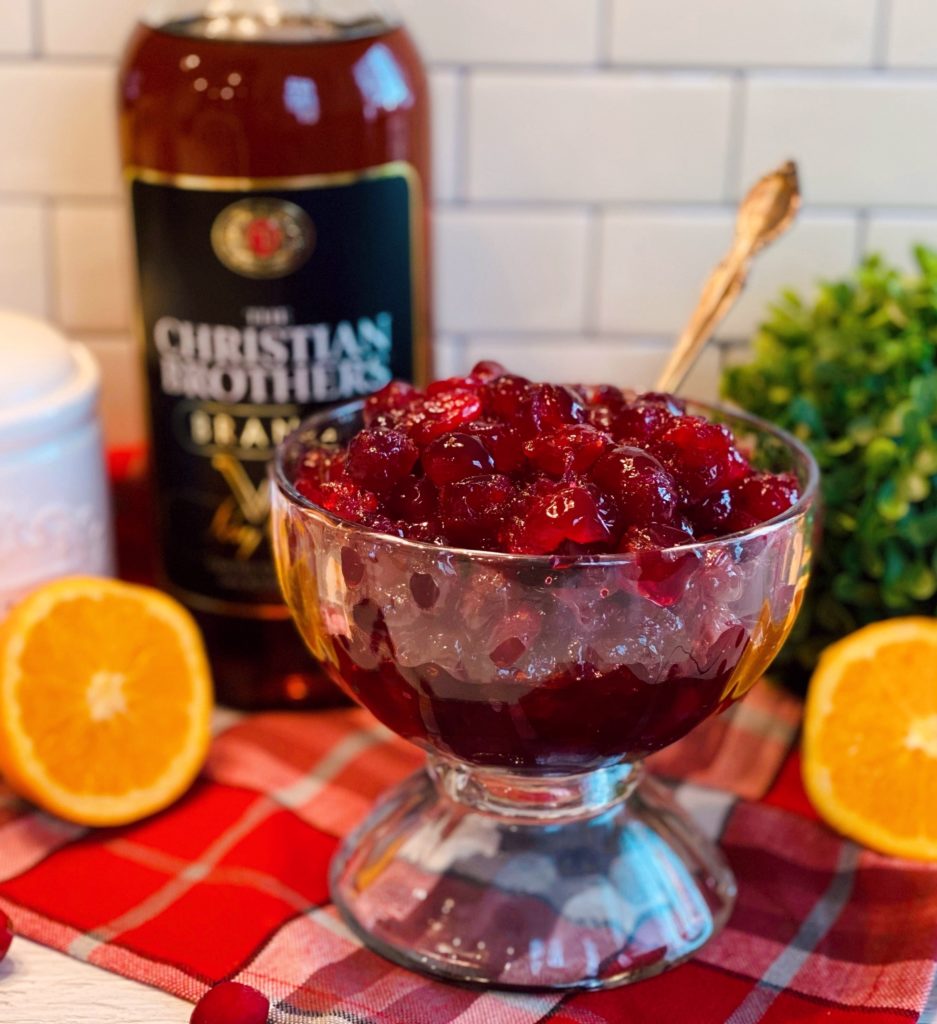 Bowl full of Baked Brandied Cranberries on a plaid table cloth with a bottle of brandy in the background.