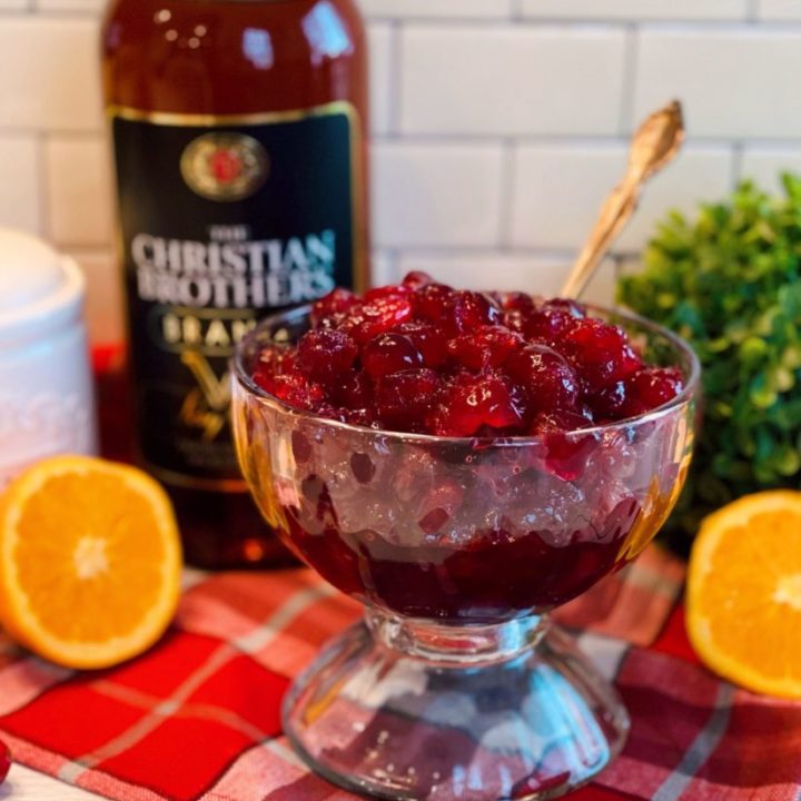 Bowl full of Baked Brandied Cranberries on a plaid table cloth with a bottle of brandy in the background.