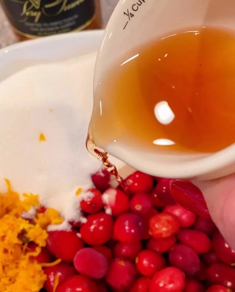 Measuring cup pouring brandy into the baking dish with cranberries, sugar, and orange zest.