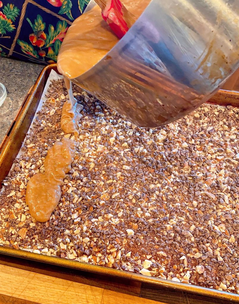 Pouring hot toffee over chocolate and nuts on baking sheet.