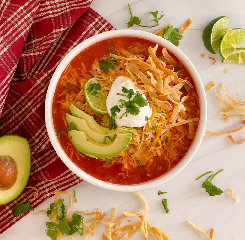 Bowl full of Chicken Tortilla Soup with fresh sliced avocado, chips, and cheese on a white counter top in a white bowl.
