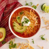 Bowl full of Chicken Tortilla Soup with fresh sliced avocado, chips, and cheese on a white counter top in a white bowl.