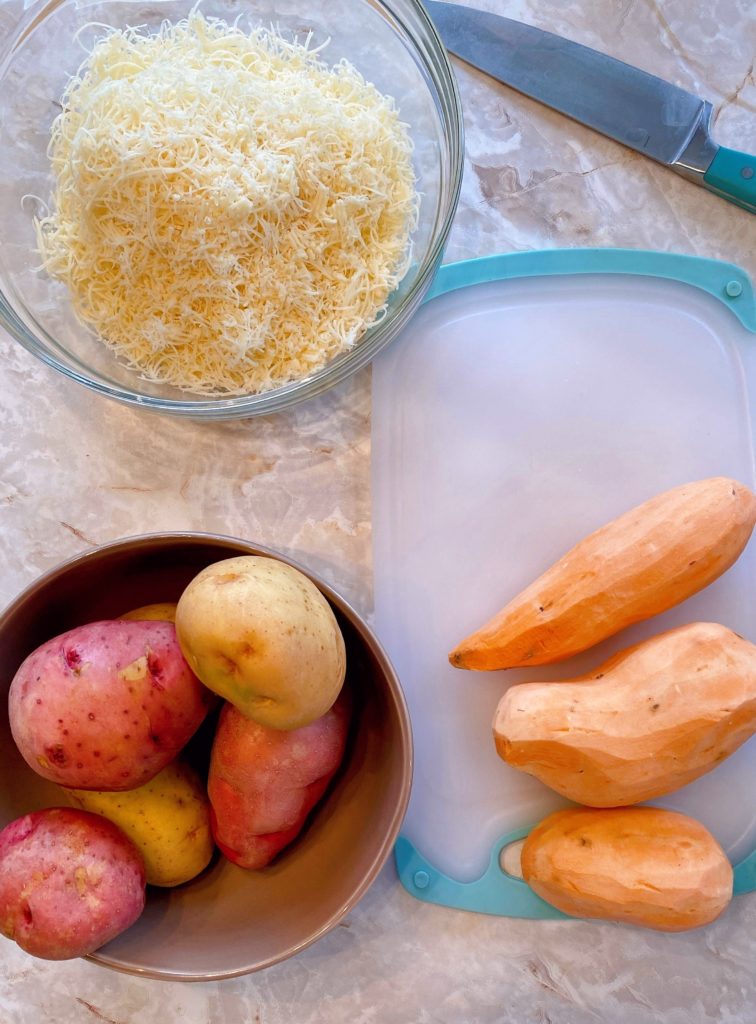 Ingredients laid on the table for Potato Gratin. Red potatoes, yukon potatoes and peeled sweet potatoes on a cutting board. Bowl with grated swiss cheese. 