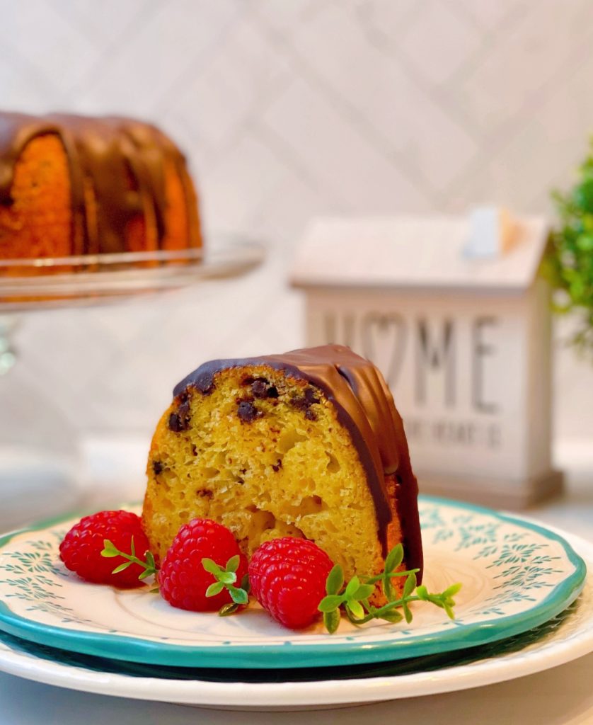 Slice of Vanilla Chocolate Chip Cake with raspberries and whole cake in the background.