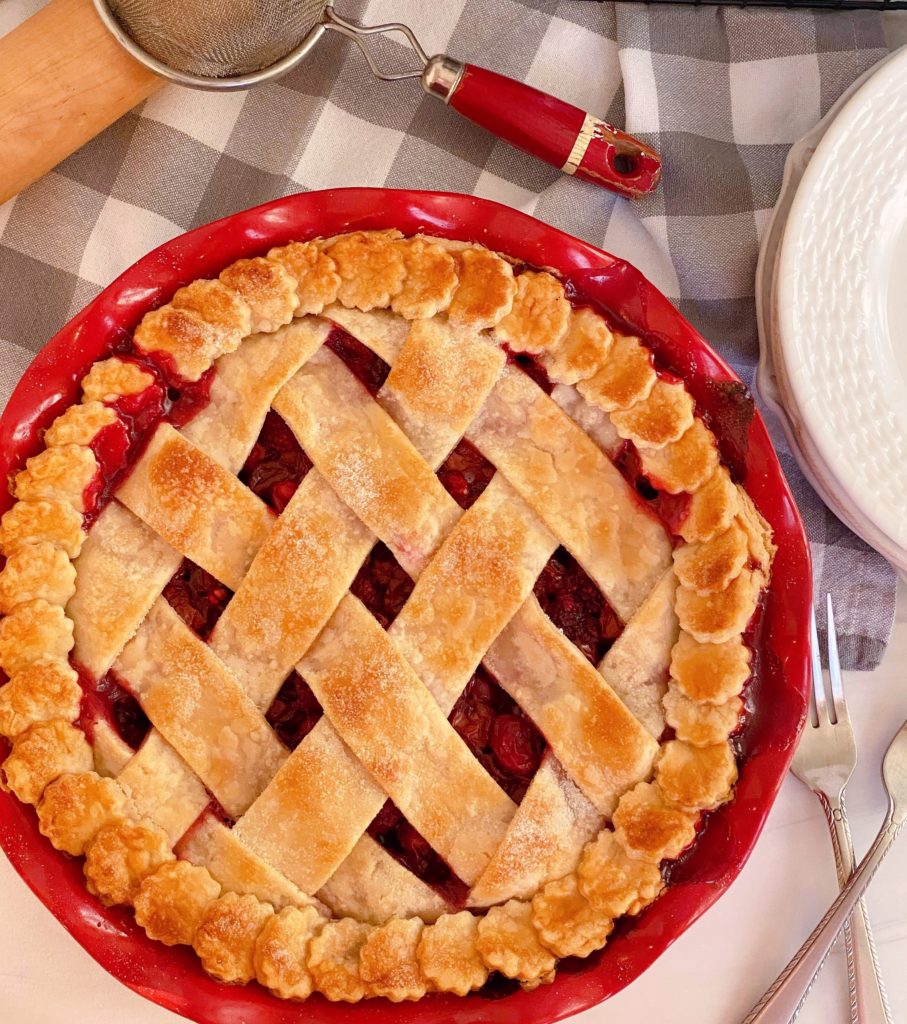 Baked Cherry Cran-raspberry pie on a checked grey cloth with a rolling pin and sifter.