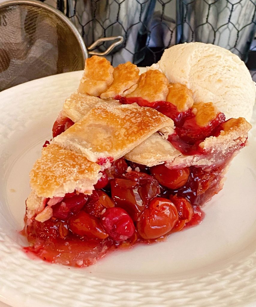 A slice of Cherry Cran-raspberry Pie on a white plate with a scoop of vanilla ice cream.