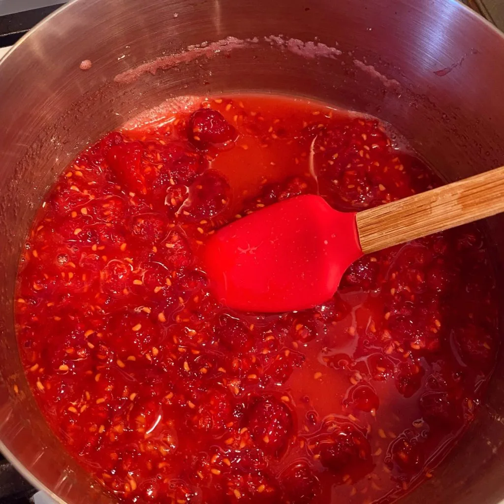Small spatula stirring raspberry sauce in stainless steel pan.