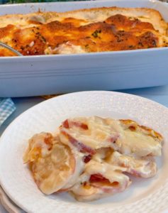 Cheesy Au Gratin Potatoes on a white plate with a casserole dish filled with Creamy Au Gratin potatoes in a casserole.