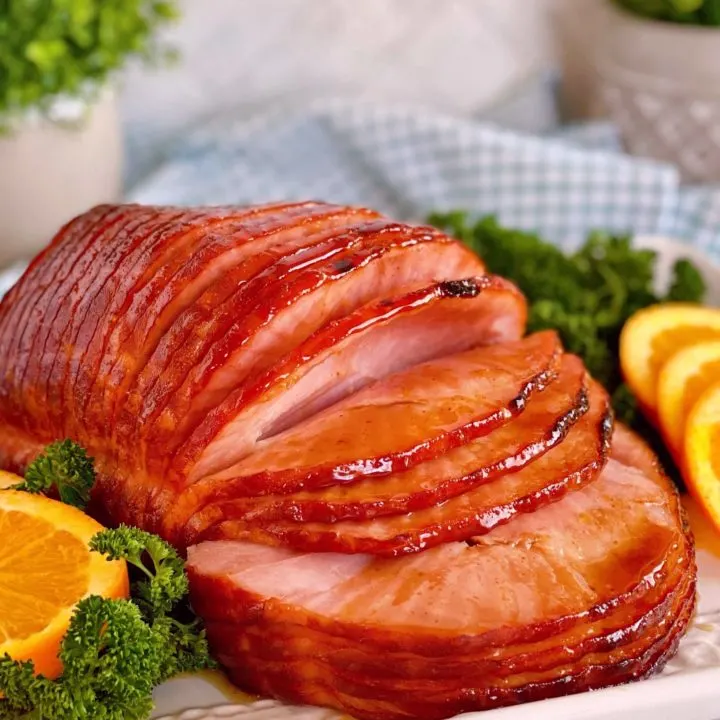 Whole Dr Pepper Glazed Spiral Ham on a serving platter surrounded by parsley and sliced oranges.