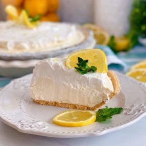 One slice lemon cream pie on a white plate with lemon slices and mint with the whole pie in the background.