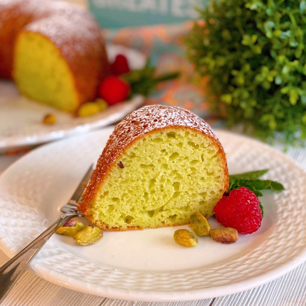 Pistachio Sour Cream Cake slice on a white plate with  the bundt cake in the back ground.