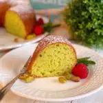 Pistachio Sour Cream Cake slice on a white plate with the bundt cake in the back ground.