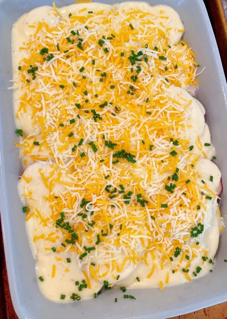 Au Gratin Potatoes ready for the oven with cheese sauce, grated cheese, and chopped chives.
