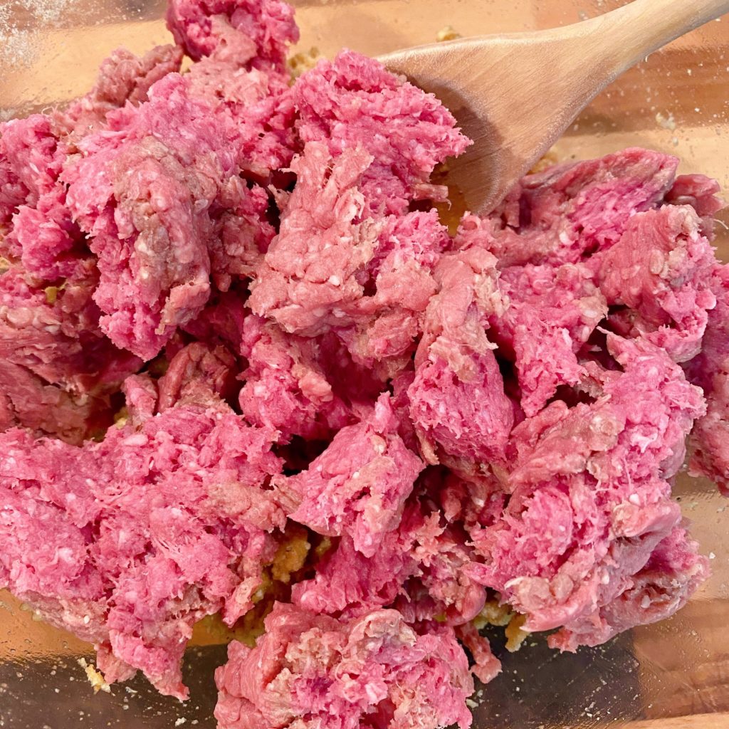 Crumbled Ground Beef over meatball ingredients in a large glass bowl.