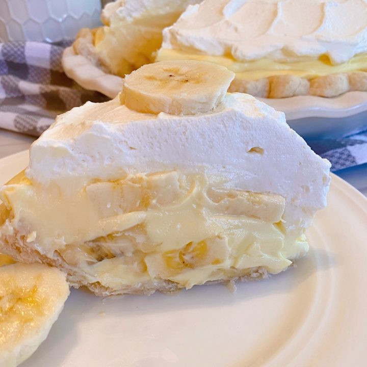Slice of Banana Cream Pie on a white dessert plate with whole pie in the background.