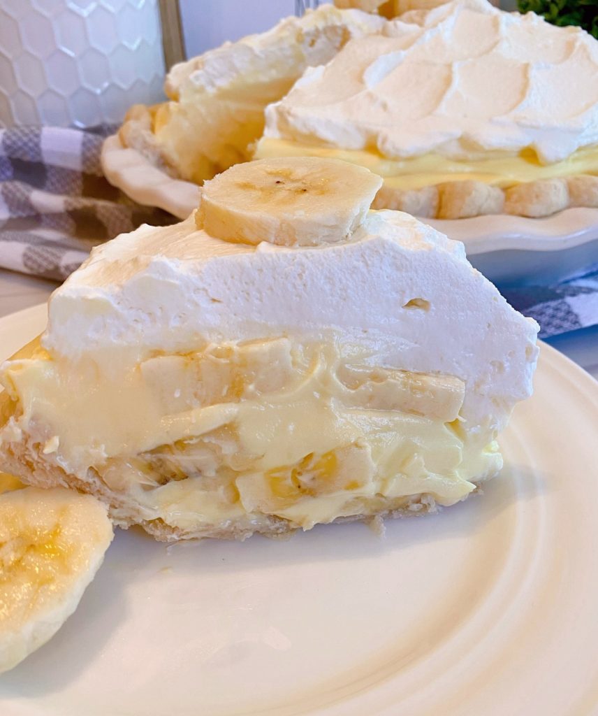 Slice of Banana Cream Pie on a white dessert plate with whole pie in the background.