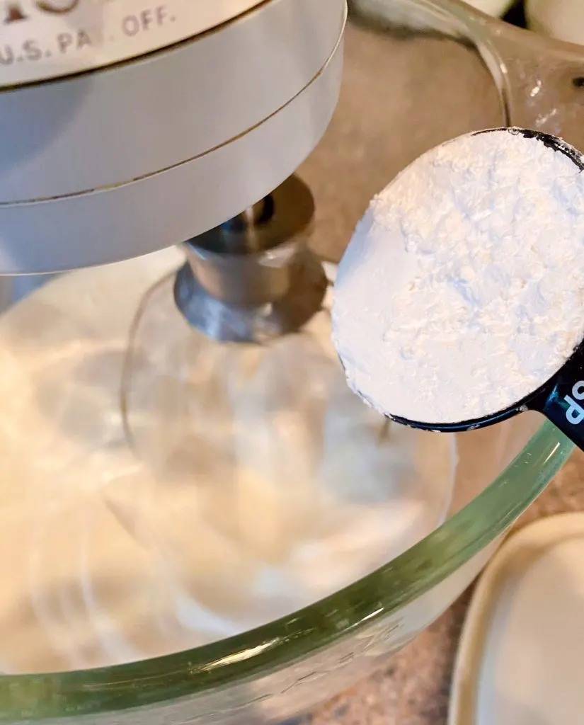 Adding Confectioners sugar to whipping cream in mixing bowl of mixer.