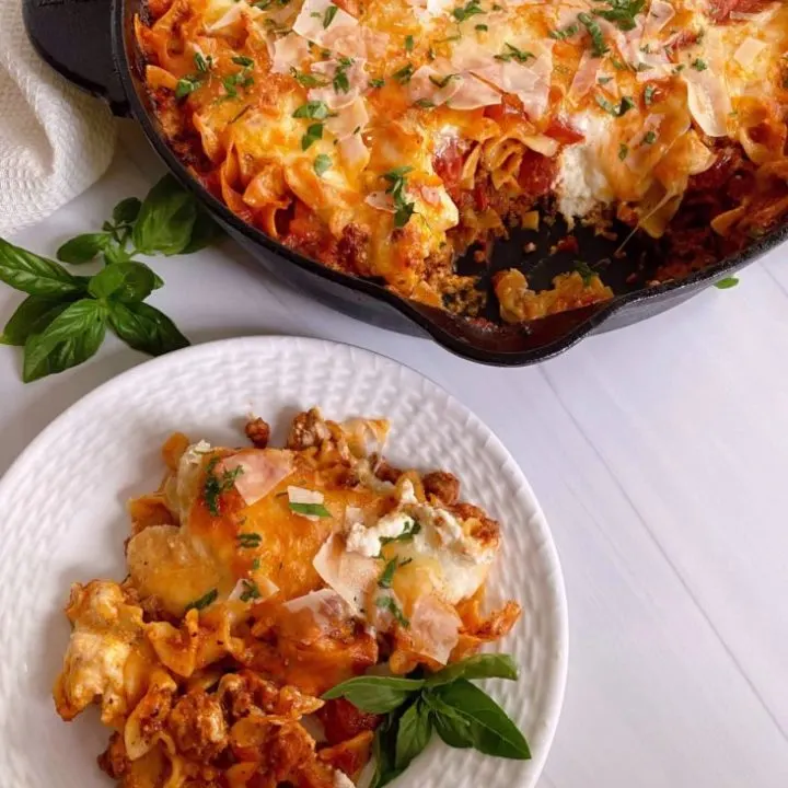 Skillet Lasagna in the skillet with a serving on a white plate on the counter top.