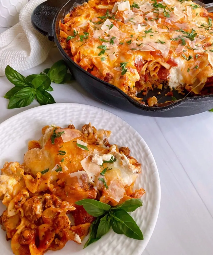 Skillet Lasagna in the skillet with a dinner plate with one serving on it garnished with fresh basil.