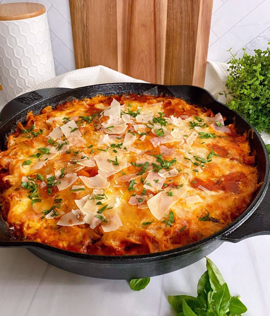 Cast Iron Skillet filled with One Pot Skillet Lasagna on a counter top with basil around it and a cutting board in the background.