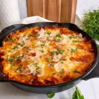 Cast Iron Skillet filled with One Pot Skillet Lasagna on a counter top with basil around it and a cutting board in the background.