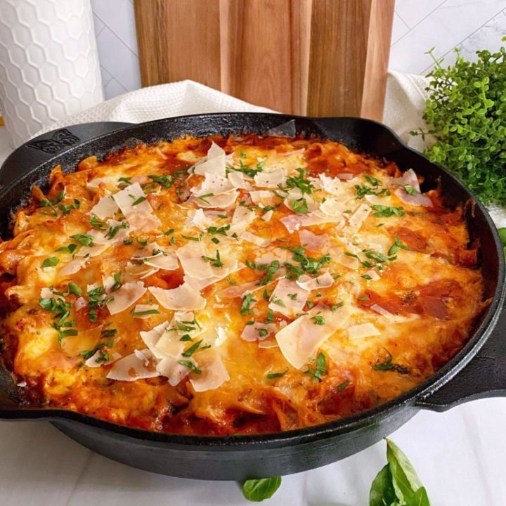 Cast Iron Skillet filled with One Pot Skillet Lasagna on a counter top with basil around it and a cutting board in the background.