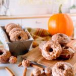 Pumpkin Spice Donuts both glazed and cinnamon sugar coated on a cutting board and in a loaf pan with a pumpkin in the background.