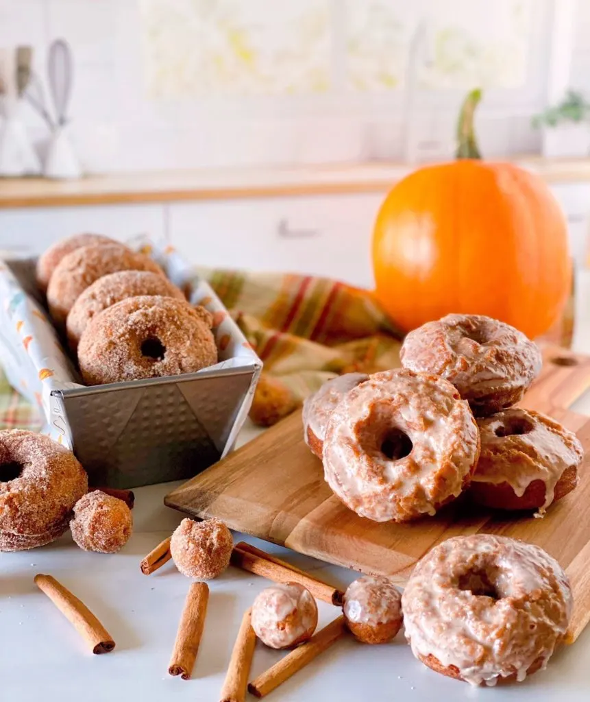 Pumpkin Spice Donuts both glazed and cinnamon sugar coated on a cutting board and in a loaf pan with a pumpkin in the background.