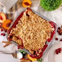 Cranberry Peach Cobbler in a baking dish surrounded by fresh peaches and cranberries on a white table.