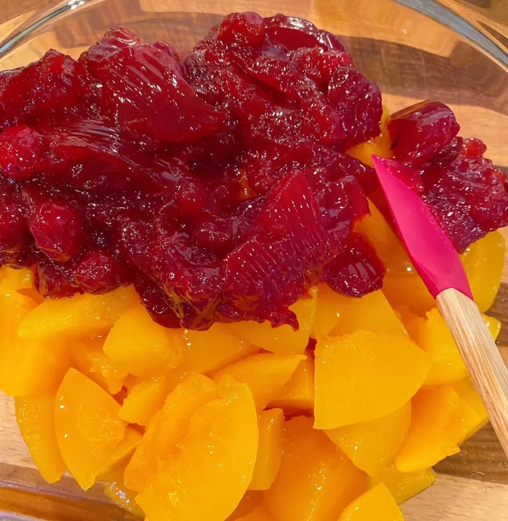 Mixing bowl with peaches and cranberry sauce being mixed together with red spatula.