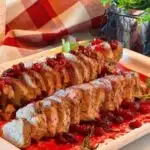 White serving platter filled with sliced Roasted Pork Tenderloin with cranberry sauce and surrounded by parsley and rosemary garnish.