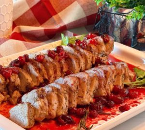 White serving platter filled with sliced Roasted Pork Tenderloin with cranberry sauce and surrounded by parsley and rosemary garnish.