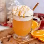 Mug of slow cooker Spiced Apple Cider with orange slices surrounding it, topped with whipped cream, caramel drizzle, and a cinnamon stick.