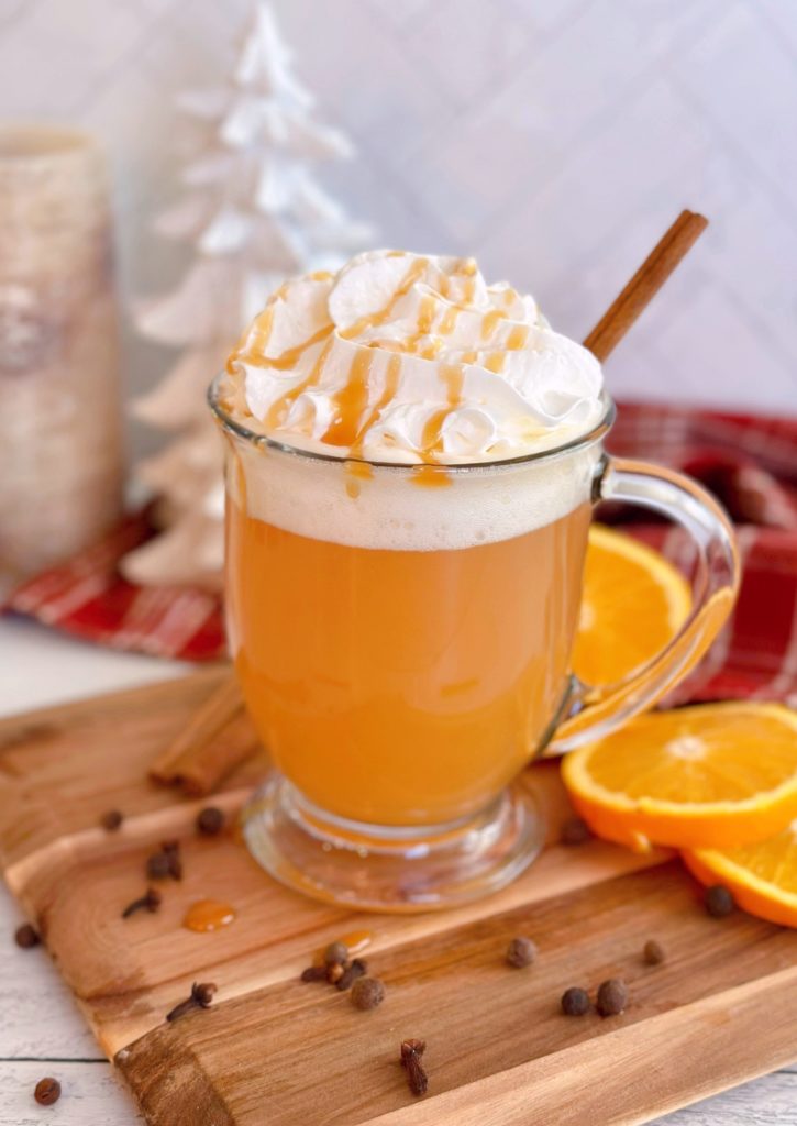 Mug of slow cooker Spiced Apple Cider with orange slices surrounding it, topped with whipped cream, caramel drizzle, and a cinnamon stick.