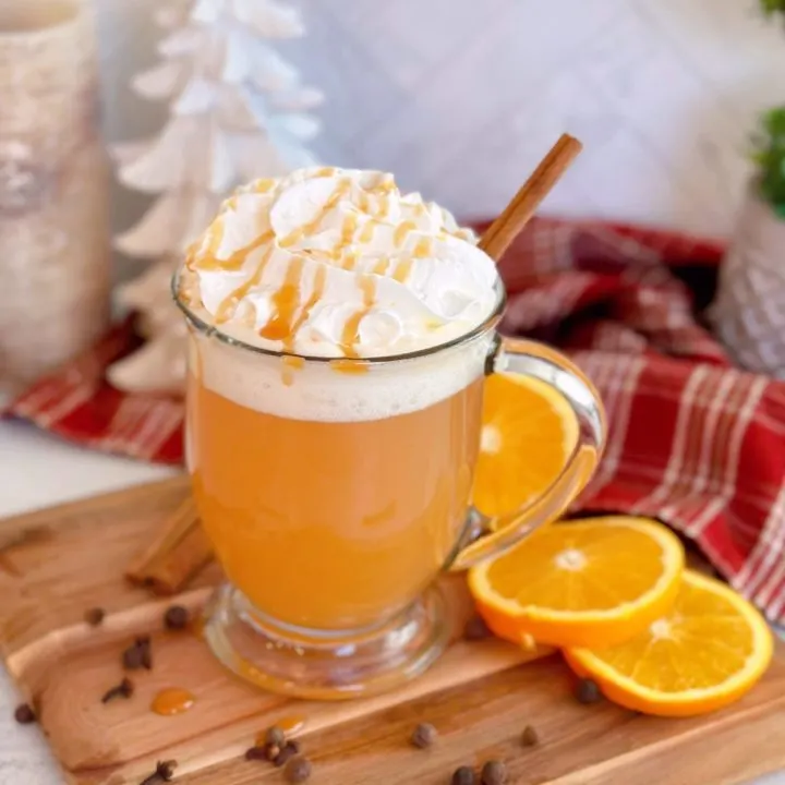 Glass of Old Fashioned Spiced Apple Cider on a cutting board with orange slices surrounding it. Topped with Whipped Cream and a caramel drizzle.