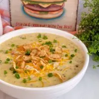 Creamy Cheeseburger Soup in a white bowl on a marble counter top with a photo of burger in the background. Topped with green onions and fried crunchy onions.