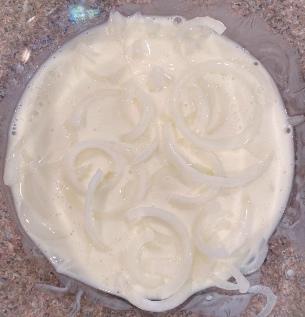 Thinly sliced onion straws soaking in buttermilk in a large bowl.