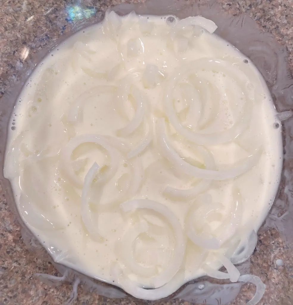 Thinly sliced onion straws soaking in buttermilk in a large bowl.