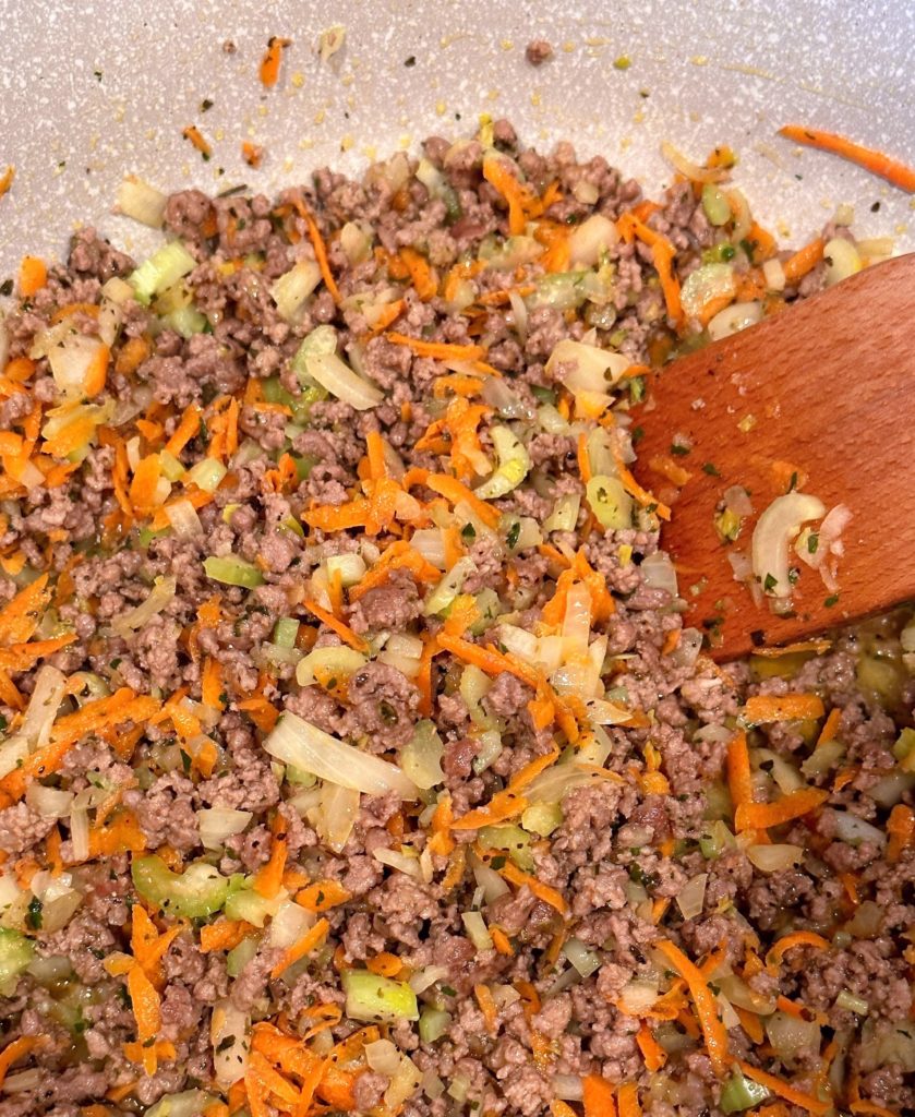 Adding vegetables and seasonings with ground beef in large pot on the stove.
