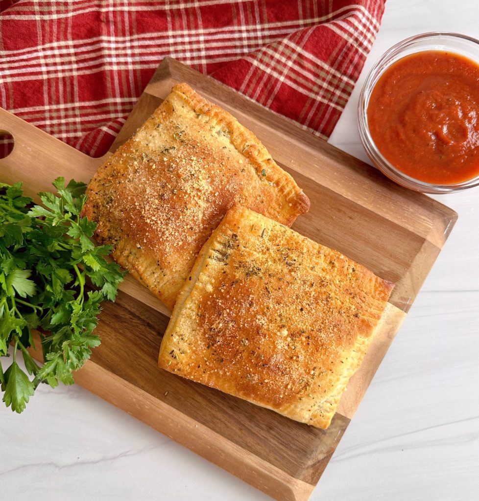 Air Fryer Pepperoni Calzones on a wooden board and a cup of marinara sauce.
