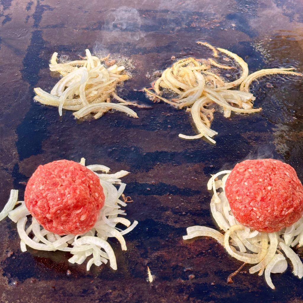 Onions down on a hot griddle surface with two meat balls on top of two of the piles.