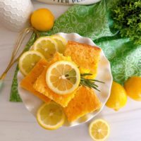 Overhead shot of Lemon Jello Cake cut into squares and piled high on a white cake plate with lemon slices.