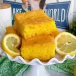 Squares of Lemon Cake piled high on a white cake plate with fresh lemon slices.