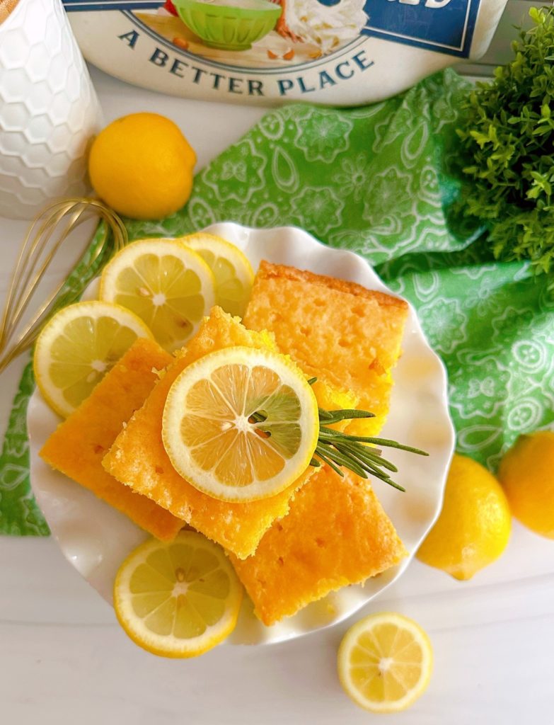 Overhead photo of slices of lemon poke cake on a white plate with fresh slices of lemon and a sprig of rosemary.