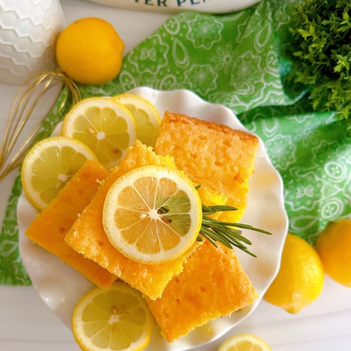 Overhead photo of slices of lemon poke cake on a white plate with fresh slices of lemon and a sprig of rosemary.