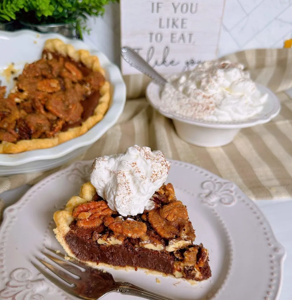 Chocolate Pecan Chess Pie sliced on a white plate with a big dollop of whipped cream and the whole pie in the background.