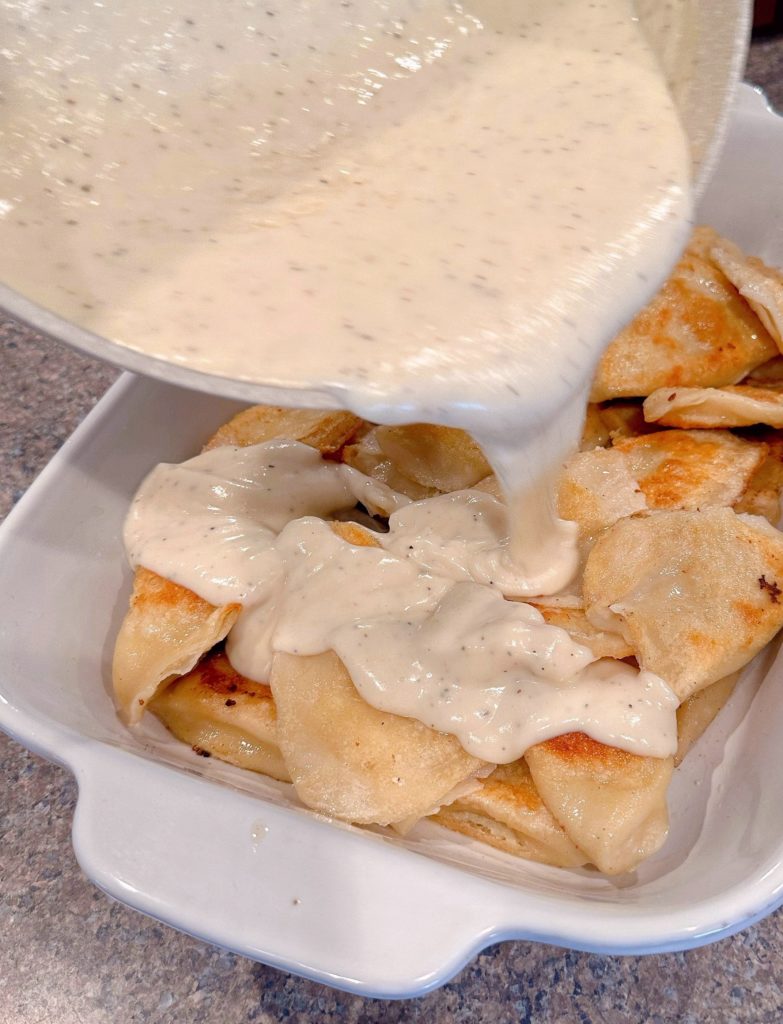 Pouring cream sauce over cooked pierogies in baking dish.