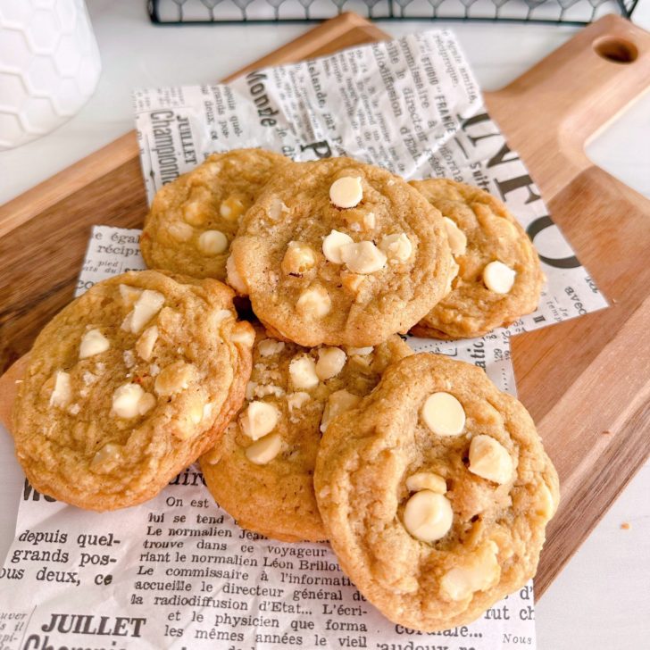 White Chocolate Chip Macadamia Nut cookies in a pile on a cutting board with news paper print.